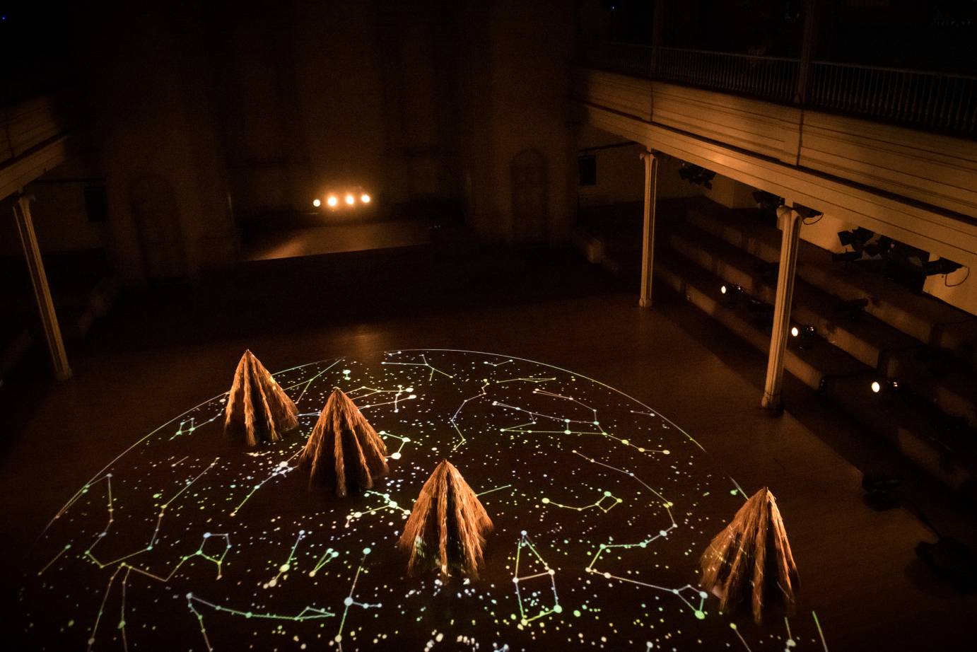 Four dancers with raffia cones covering them stand on a floor illuminated with a projection of stars and constellations